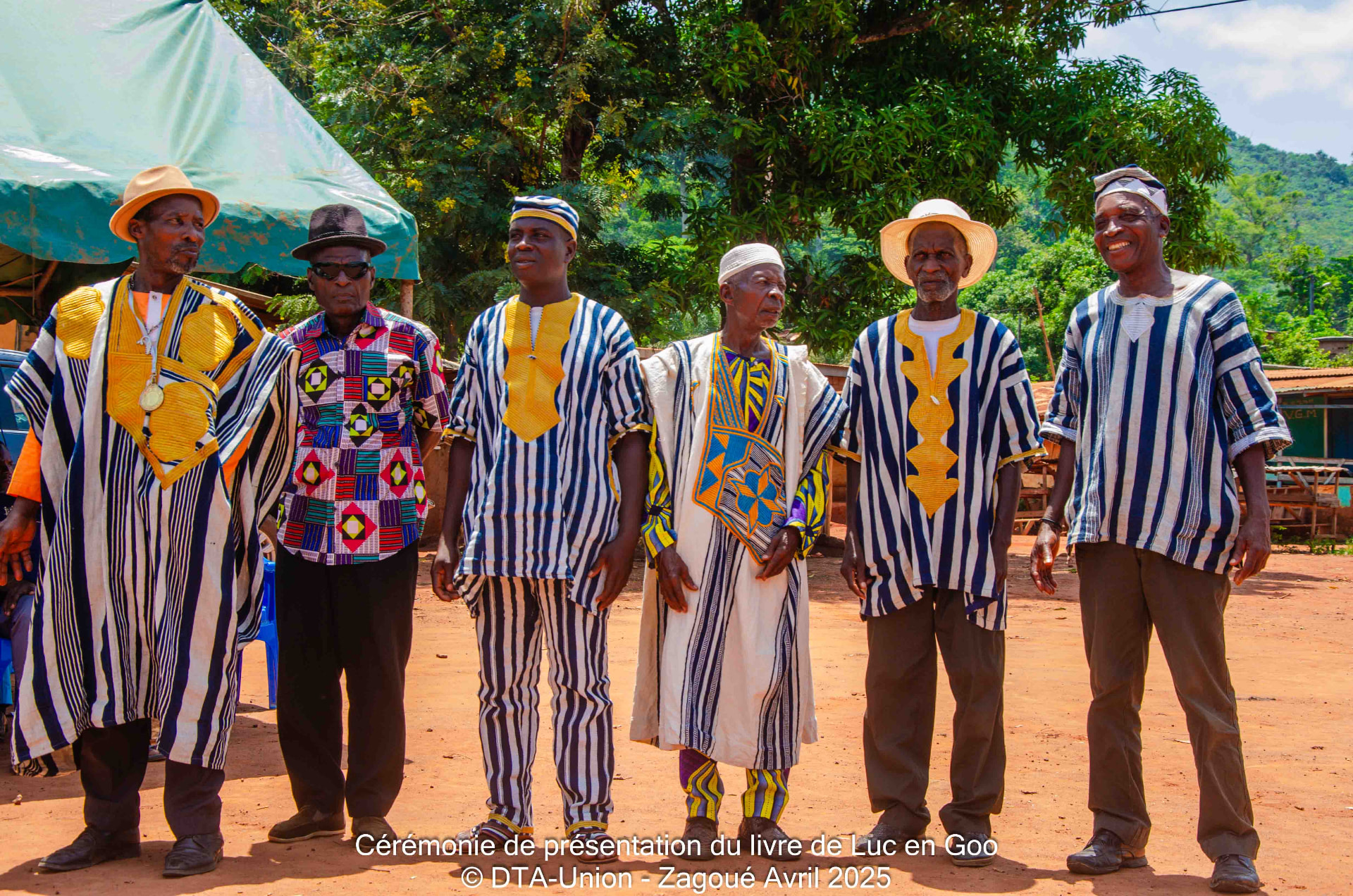 Six anciens de Zagoué en pagne tissé alignés sur la place du village, avril 2025